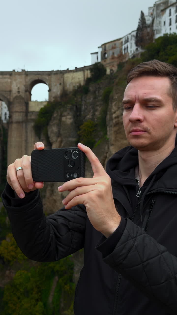 Mand ved kløftbro fotograferer landskab med smartphone fra klippekant, historisk stenbue.