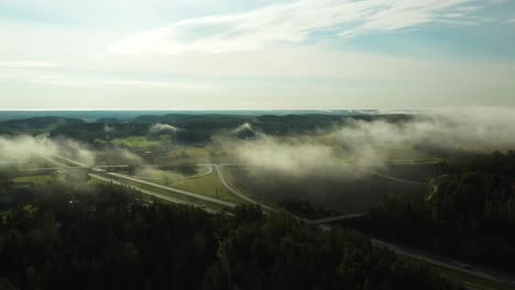 forward moving aerial view of morning fog floating over a 4-lane highway with cars and trucks. beatiful forests and fields below partly clouded sky.