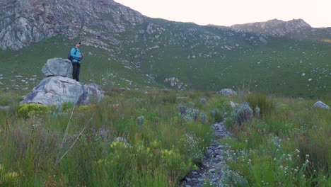 man on hike, takes break on boulder drinking water with massive sandstone mountain in background