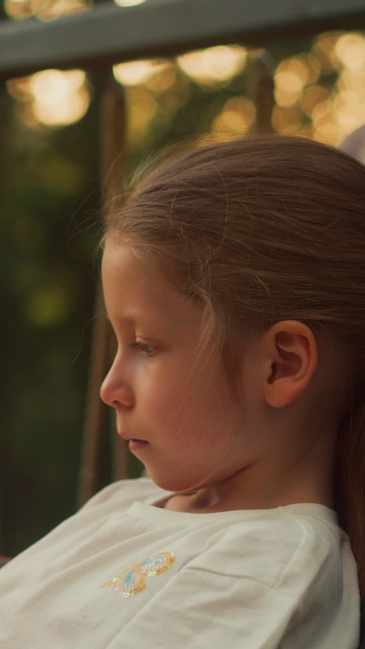 niña en edad preescolar pasa el tiempo jugando a un juego móvil en la terraza. niño tranquilo usa un teléfono inteligente reclinado en un cómodo sillón contra el bosque salvaje de verano