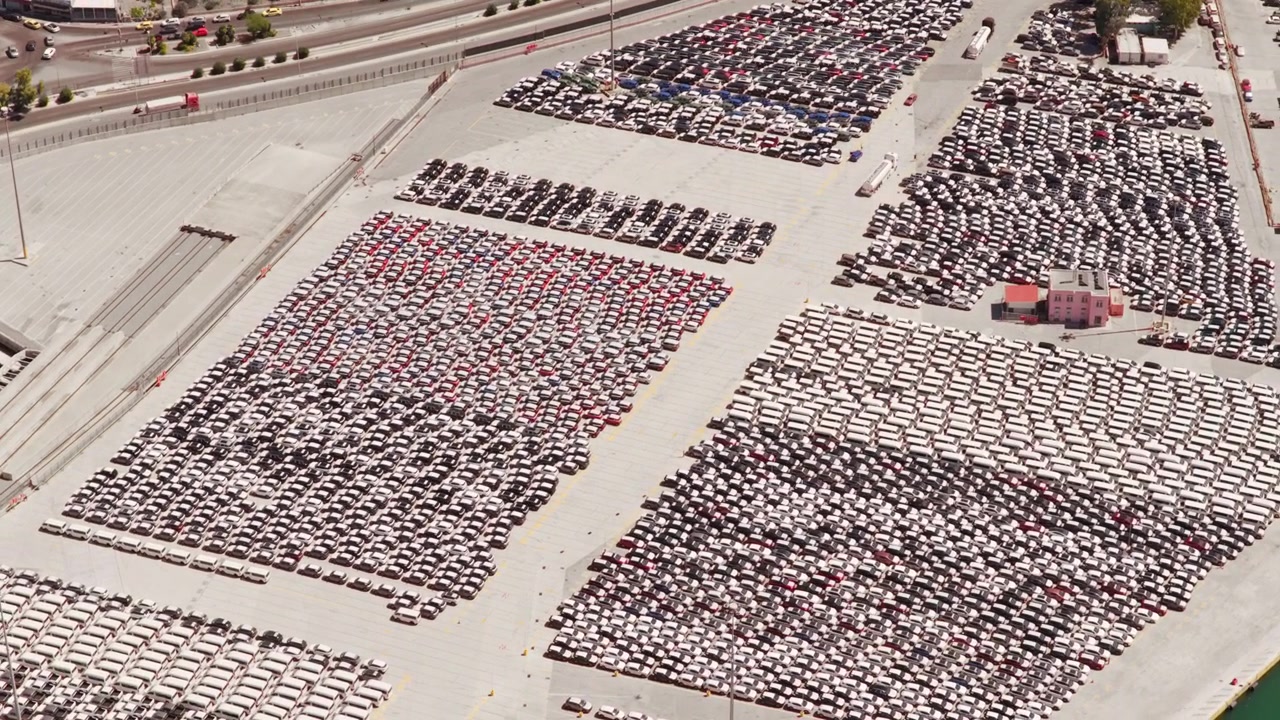 High-angle view of a vast parking lot in an industrial port, packed with thousands of new cars sorted by color and ready for shipping, distribution, and export to the global market