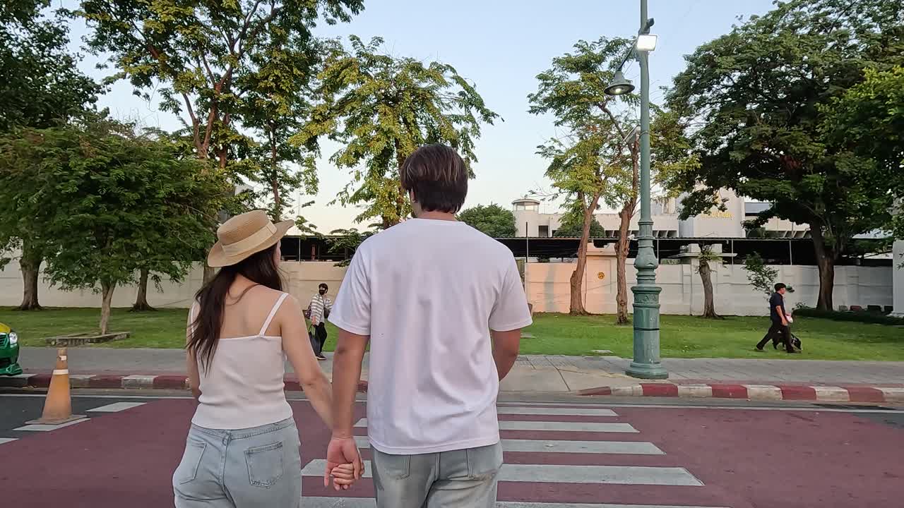 Rear view of couple holding hands walking across city park crosswalk at sunset