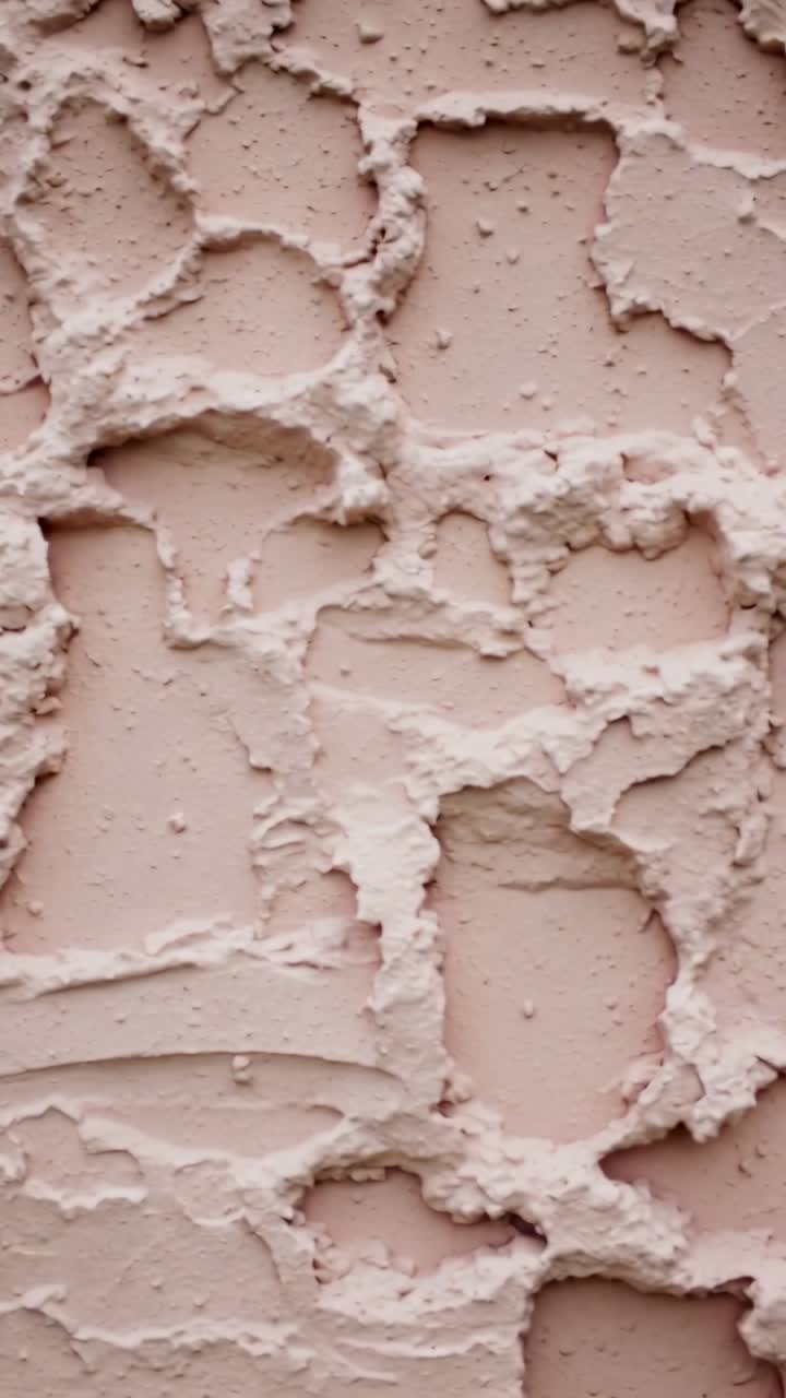 Vertical video: Moving phone filming closeup stucco wall indoors, showing muted pink-beige specks