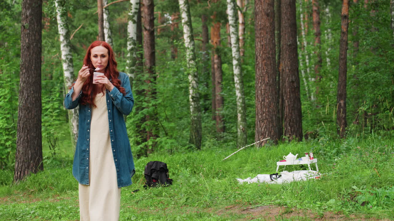 여자가 숲 속 빈터에서 아이스크림 컵을 들고, 공원에서 편안한 여름 가족 나들이를 즐기는 동안 피크닉 매트에 앉아 있는 딸을 사랑스럽게 바라보며 걷고 있어요. 주변은 자연으로 둘러싸여 있네요.