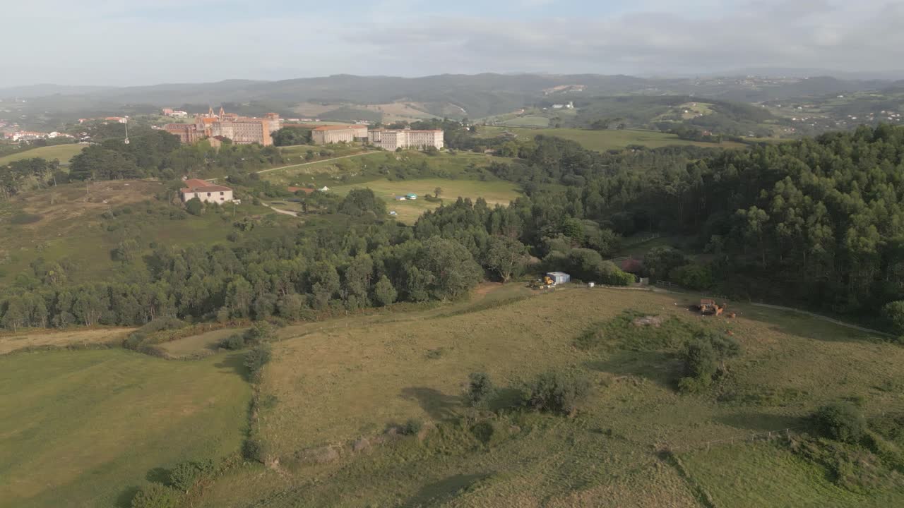 Stationary drone shot with a left yaw over the outer fields of Comillas, showing green meadows, tree-filled estates, the iconic university behind, and the coastline with its beaches