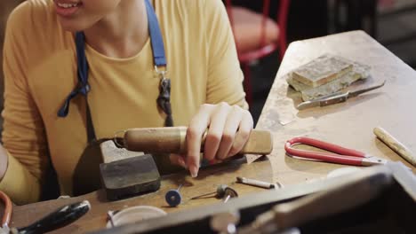 happy biracial female worker at table shaping ring with handcraft tools in studio in slow motion