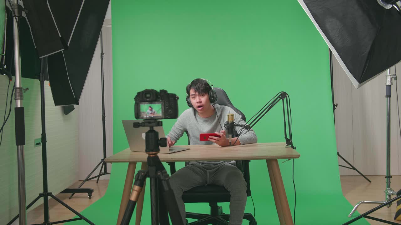 A Camera Recording Asian Man Gamer With Headphone Reading Comment On Computer While Playing Game On Mobile Phone On Green Screen With Professional Light Equipment