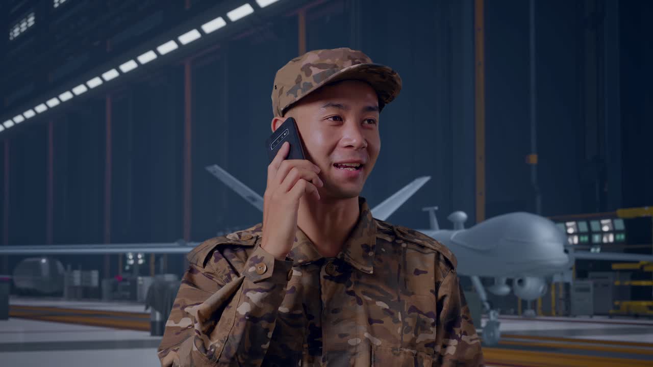 Close Up Of Asian Man Soldier Talking On Smartphone And Looking Around While Standing With Modern Military Drone In Maintenance Hangar