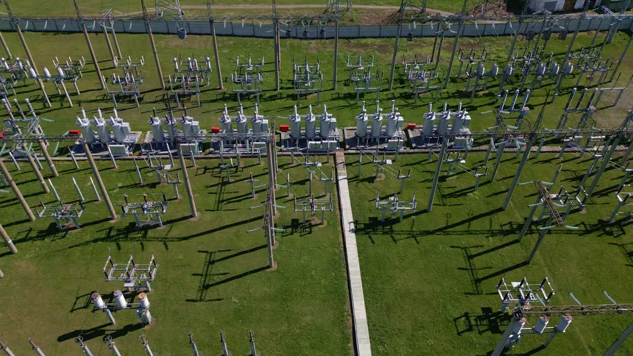 Luftaufnahme eines weitläufigen Umspannwerks. Grünes Gras umgibt hoch aufragende elektrische Anlagen. Bauwerke werfen lange Schatten unter hellem Sonnenlicht. In dieser Szene sind keine Personen oder Tiere zu sehen.