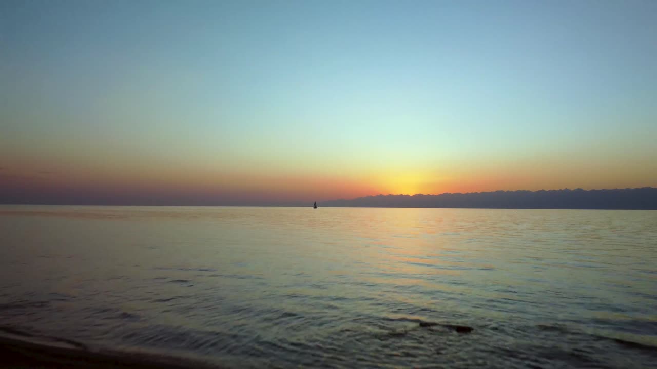 드론이 장엄한 산 뒤로 해가 질 때 물 위를 낮게 날아가는 해변의 멋진 영상을 캡처합니다.