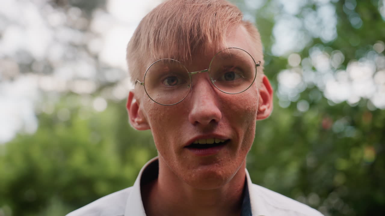 Close up view of young man in white shirt with glasses walking tired in blurred green forest background, touching face and showing signs of exhaustion in natural outdoor environment