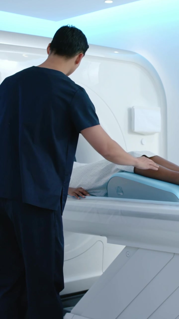 Video verticale: Tecnico in divisa blu scuro che posiziona il paziente sul tavolo della risonanza magnetica con un cuscino di supporto blu, preparando la scansione.