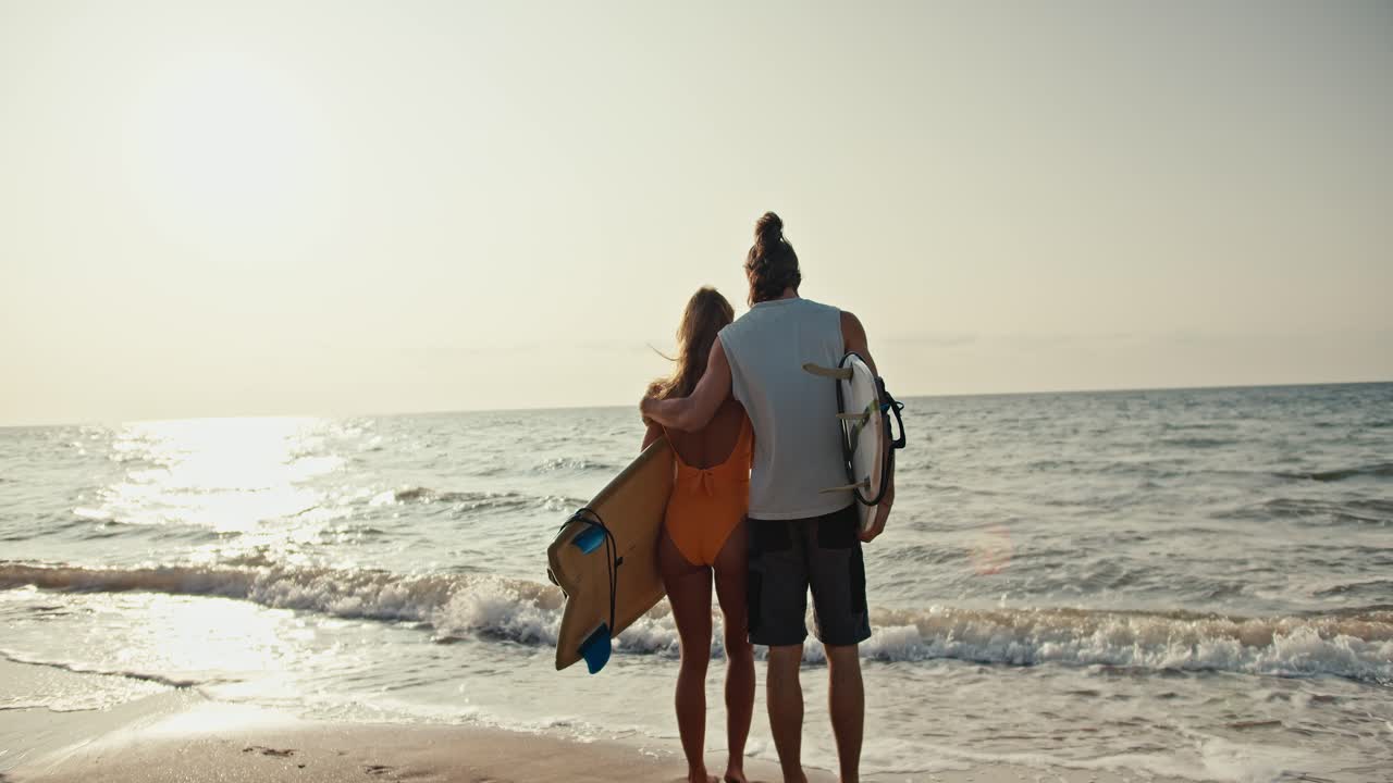 vista trasera de una pareja feliz, un hombre moreno en una camiseta blanca y su novia rubia en un traje de baño naranja sosteniendo tablas de surf de pie en la playa de arena y mirando hacia el mar al amanecer de la mañana