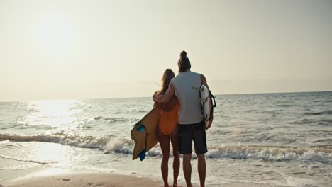 Rear-view-of-a-happy-couple,-a-brunette-man-in-a-white-tank-top-and-his-blonde-girlfriend-in-an-orange-swimsuit-holding-surfboards-standing-on-the-Sandy-Beach-and-looking-towards-the-sea-at-Sunrise-in-the-morning