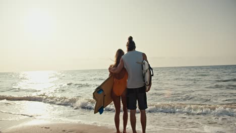 rear view of a happy couple, a brunette man in a white tank top and his blonde girlfriend in an orange swimsuit holding surfboards standing on the sandy beach and looking towards the sea at sunrise in the morning