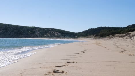 Slow-motion-shot-of-waves-crashing-on-beautiful-sandy-beach-surrounded-by-green-hills-in-South-Sardinia,-Italy