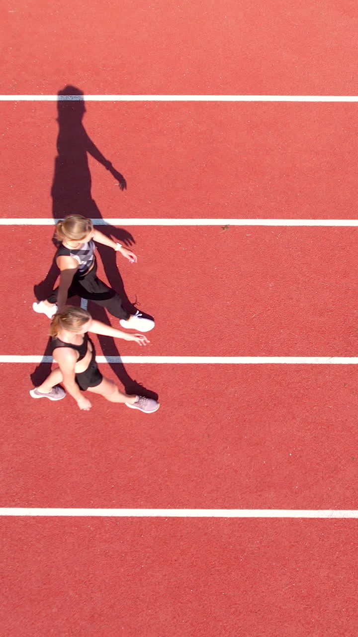 Toma aérea vertical de dos atletas femeninas caminando por los carriles blancos de una pista de atletismo roja de estadio. Ideal para contenido de fitness, deportes y estilo de vida saludable.