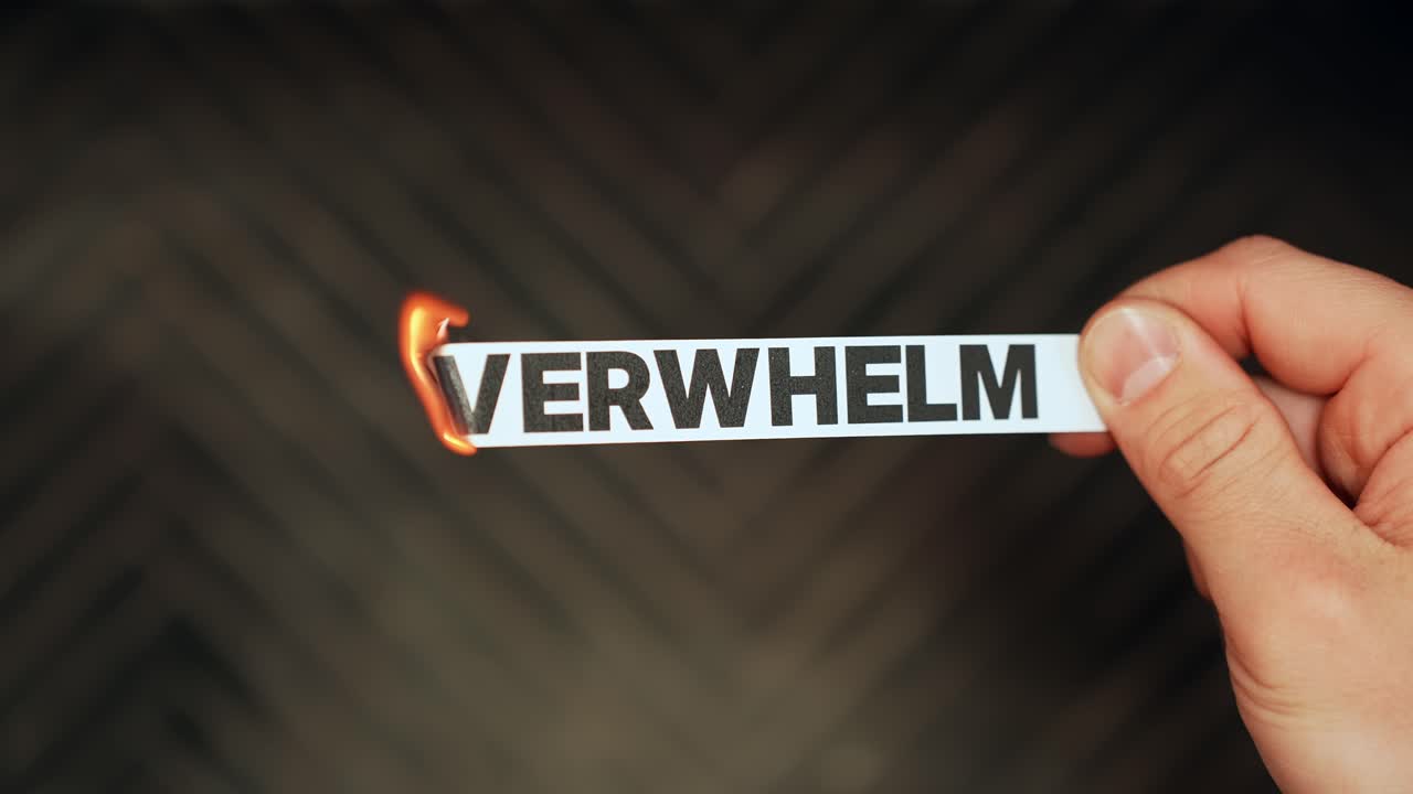 Close-up hand holds a paper strip reading “OVERWHELM” while the edge catches fire from a lighter, symbolizing stress and burnout on a dark background with copy space
