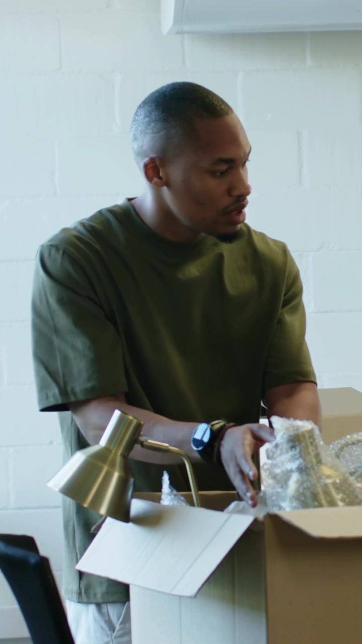 Vertical video: Leaning forward man in olive tee opening box on table inspecting wrapped gold lamp