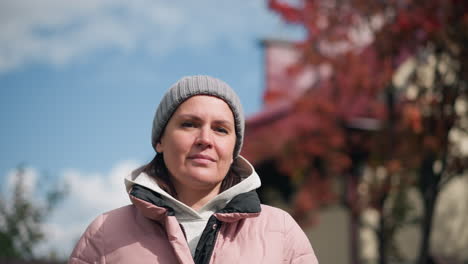 business woman in pink jacket and grey beanie, thoughtfully looking with head tilted, outdoors in autumn, posing with blurred colorful trees and house in background under clear sky