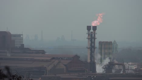 red and blue flames burning out of a chimney of a factory with more plants on the background on a cloudy grey day in the ruhr area