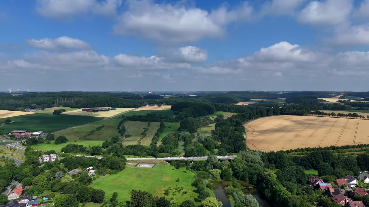 Aerial lateral weide shot of traffic on interstate road between american neighborhood and farm fields. Wind turbines in distance. BLue sky and clouds in summer. Houses and homes in small city of USA.