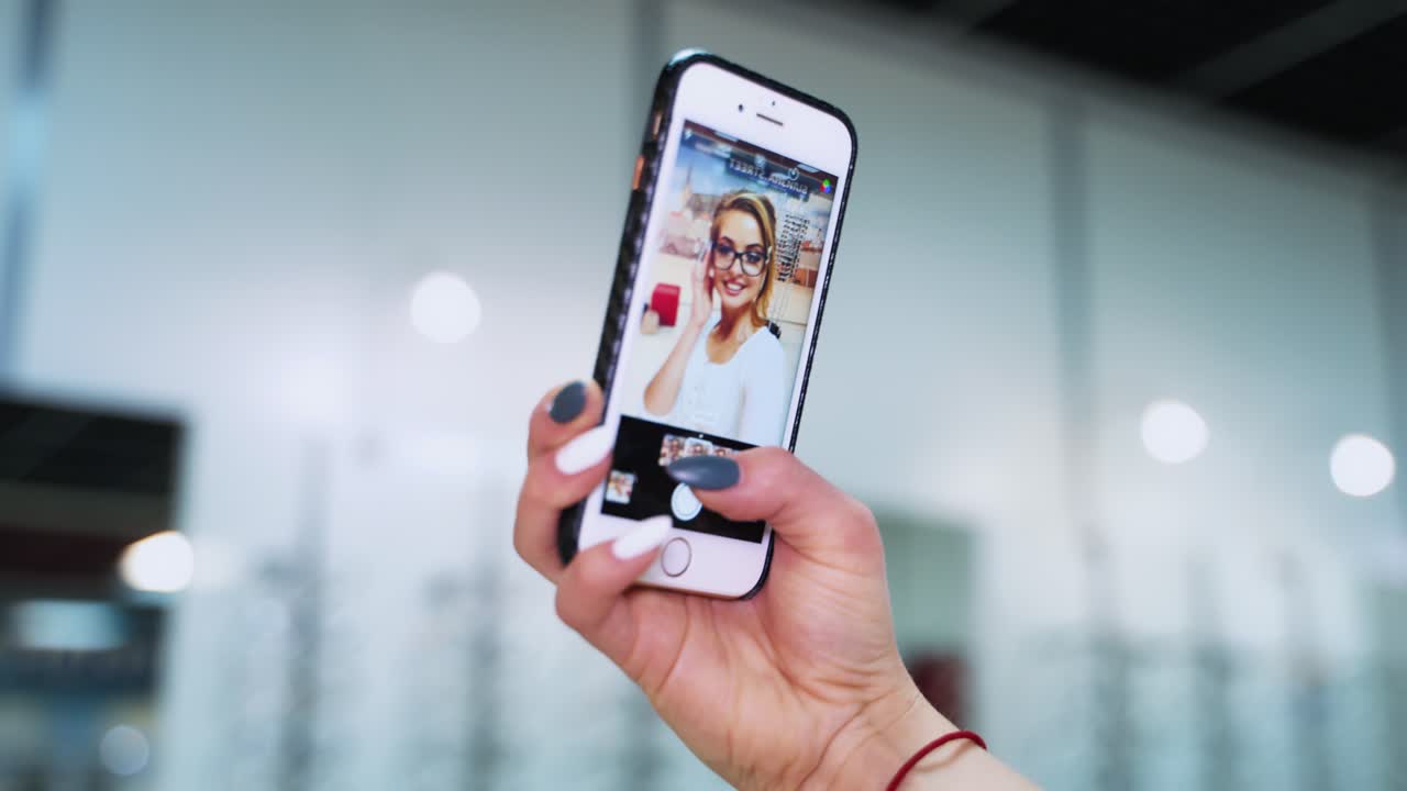 Woman looks at herself in the mobile phone. Female's hand holding phone and showing the video with a beautiful woman in eyeglasses.