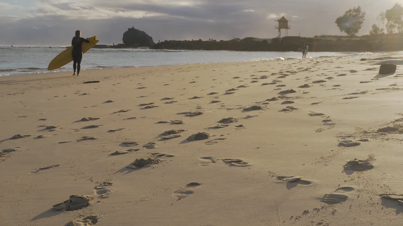 Surfista donna con tavola da surf che lascia la spiaggia dopo essersi goduta le onde al tramonto - Impronte sulla sabbia a Currumbin Beach - Gold Coast, Australia.