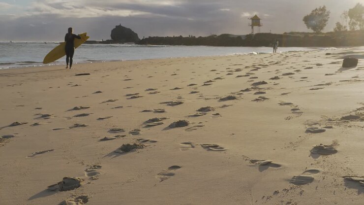 Surfista donna con tavola da surf che lascia la spiaggia dopo essersi goduta le onde al tramonto - Impronte sulla sabbia a Currumbin Beach - Gold Coast, Australia.