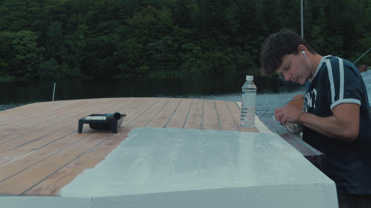 Young carpenter working on wooden boat wipes excess paint off roof