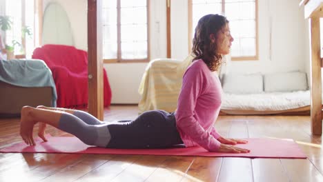 Relaxed-mixed-race-woman-practicing-yoga,-lying-on-mat-stretching-in-sunny-cottage-bedroom