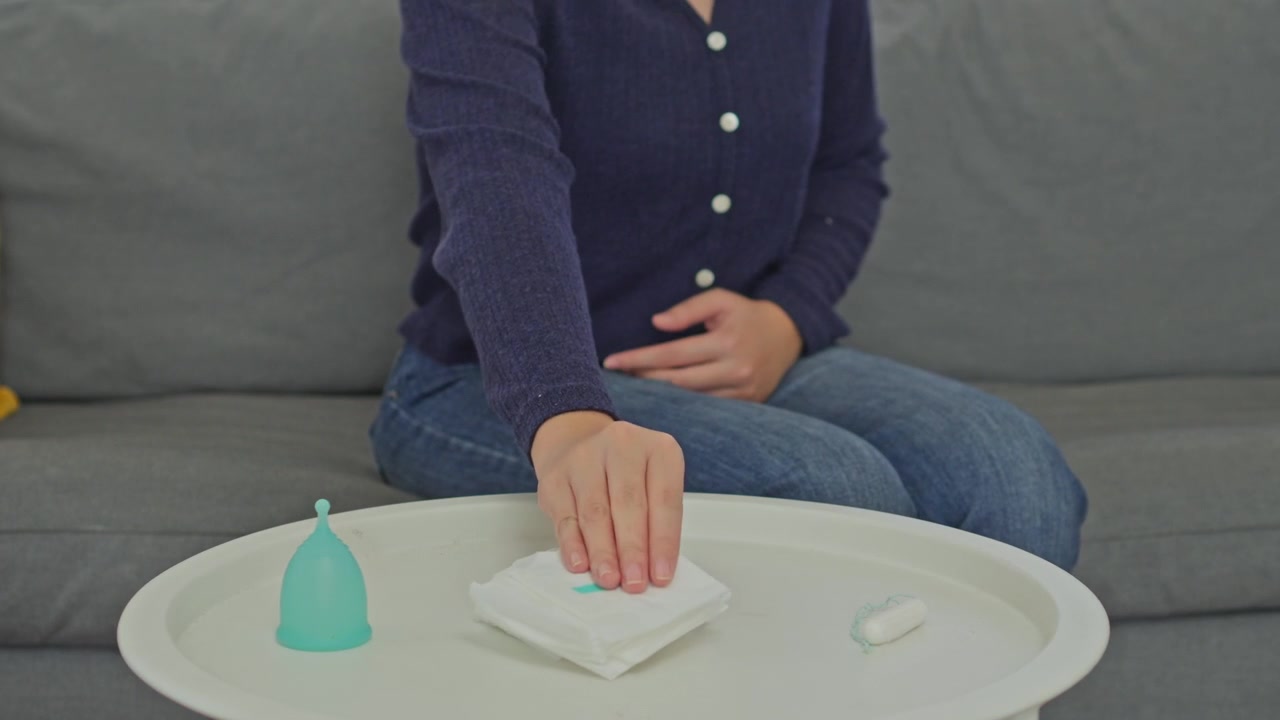 Woman experiencing period cramps reaches for sanitary pad from table with hygiene products