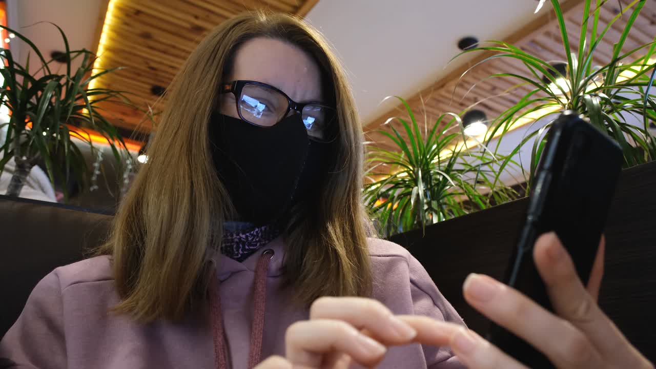 카페에서 휴대전화로 일하는 소녀 또는 젊은 여성. 카페테리아에서 얼굴에 보호 마스크를 쓴 여성이 소셜 네트워크를 스크롤합니다.