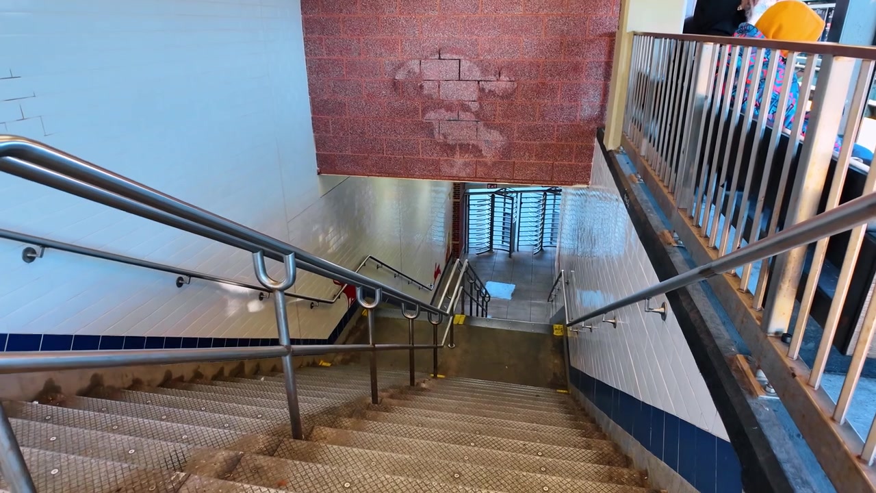 Metrostationstrappe med metalgelændere og flisebelagte vægge. Betontrin fører ned mod et drejekors inde i en offentlig transportterminal med røde murstensvægge.