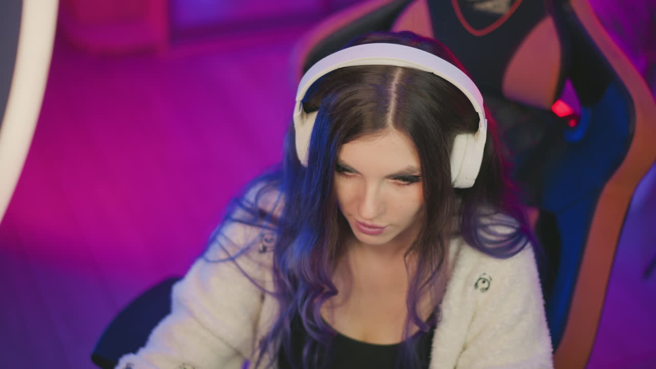top down view of female designer wearing white headphones seated in gaming chair intently working on computer in colorful setup with blurred equipment surrounding workspace