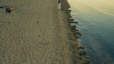 Aerial-drone-shot-of-lady-in-white-dress-at-a-lonely-beach-in-blue-hour