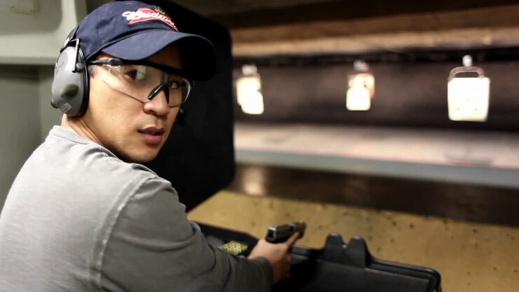 A man looks at the camera and then fires a hand gun at a target at an indoor shooting range