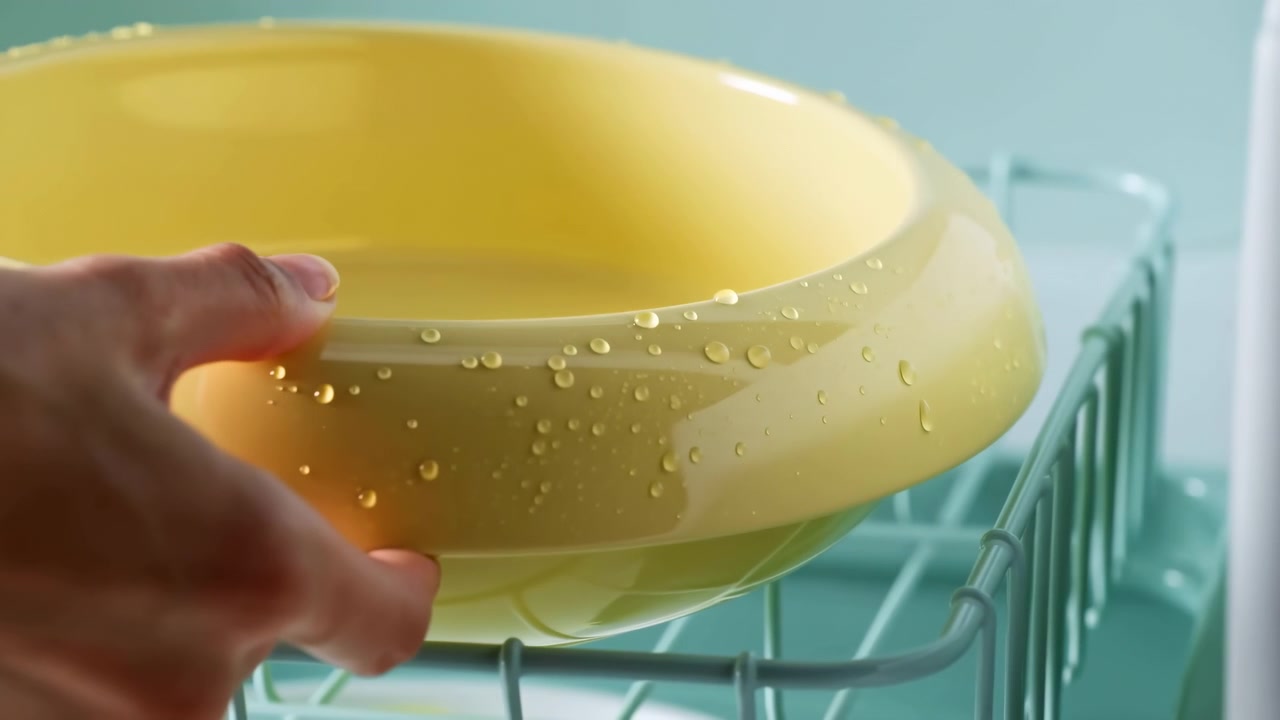 Primo piano di una mano che tiene una ciotola di plastica gialla con goccioline d'acqua sopra una griglia della lavastoviglie in una cucina turchese brillante con una morbida luce naturale.