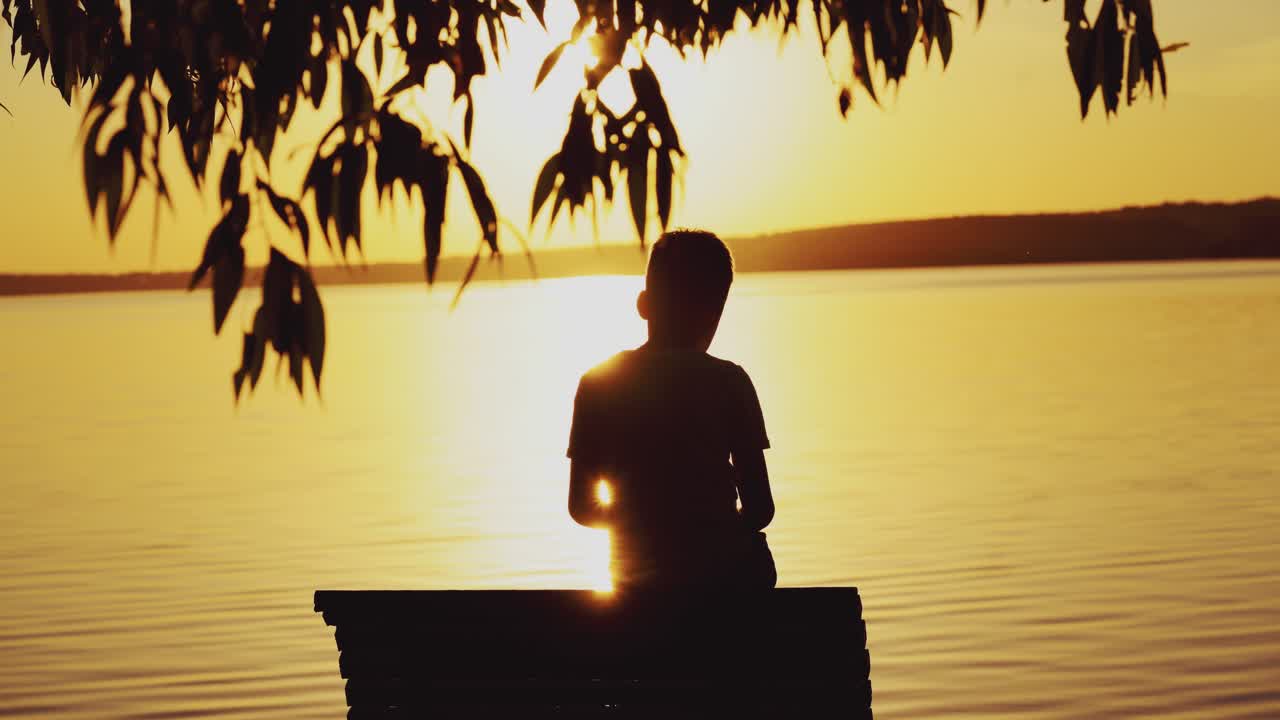 Small boy is sitting on a wooden dock at the river in the evening. Photo of a boy at sunset looking on the river and trying to find somebody or something in summer