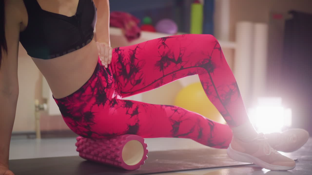 Primer plano de piernas utilizando rodillo de espuma para liberar la cadera, sujetador deportivo negro, leggings rosados estampados, esterilla de estudio interior, rodillo texturizado bajo la banda iliotibial, movimiento lento y controlado, masaje muscular, contraluz cálido, calma y concentración.