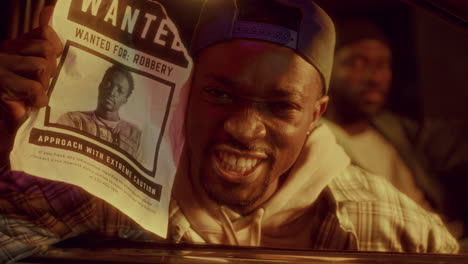 grinning black man holding wanted poster with his own photo in police lights