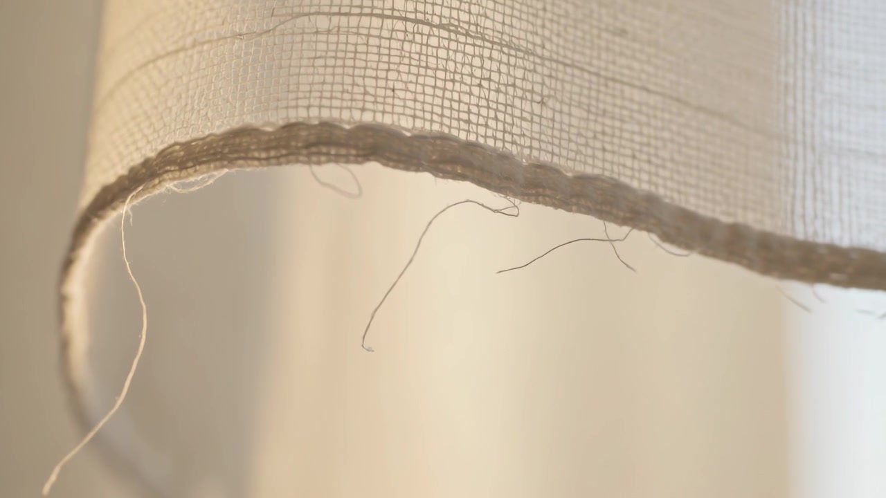 Brezza leggera che muove l'orlo di una tenda beige chiaro vicino alla finestra, mostrando il bordo cucito e i fili sfilacciati.