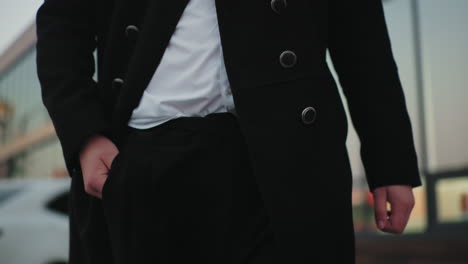 close up of man in black coat placing hand inside pocket with background featuring parked cars and building, confident, stylish individual in urban setting, exuding sophistication and professionalism