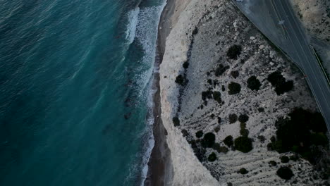 Das-Bild-Bietet-Eine-Diagonale-Luftaufnahme-Einer-Küstenszene,-Wo-Ein-Tiefblaues-Meer-Auf-Eine-Kreideweiße-Klippe-Trifft,-Die-Von-Grünem-Gebüsch-Gesäumt-Ist,-Neben-Einem-Sandstrand-Und-Einer-Straße---Zypern