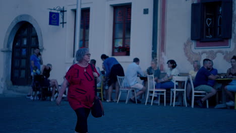 older white woman with grey hair wearing glasses and red t-shirt walking around the historical city bardejov in slovakia during a blue hour dusk in slow motion, surrounded by people