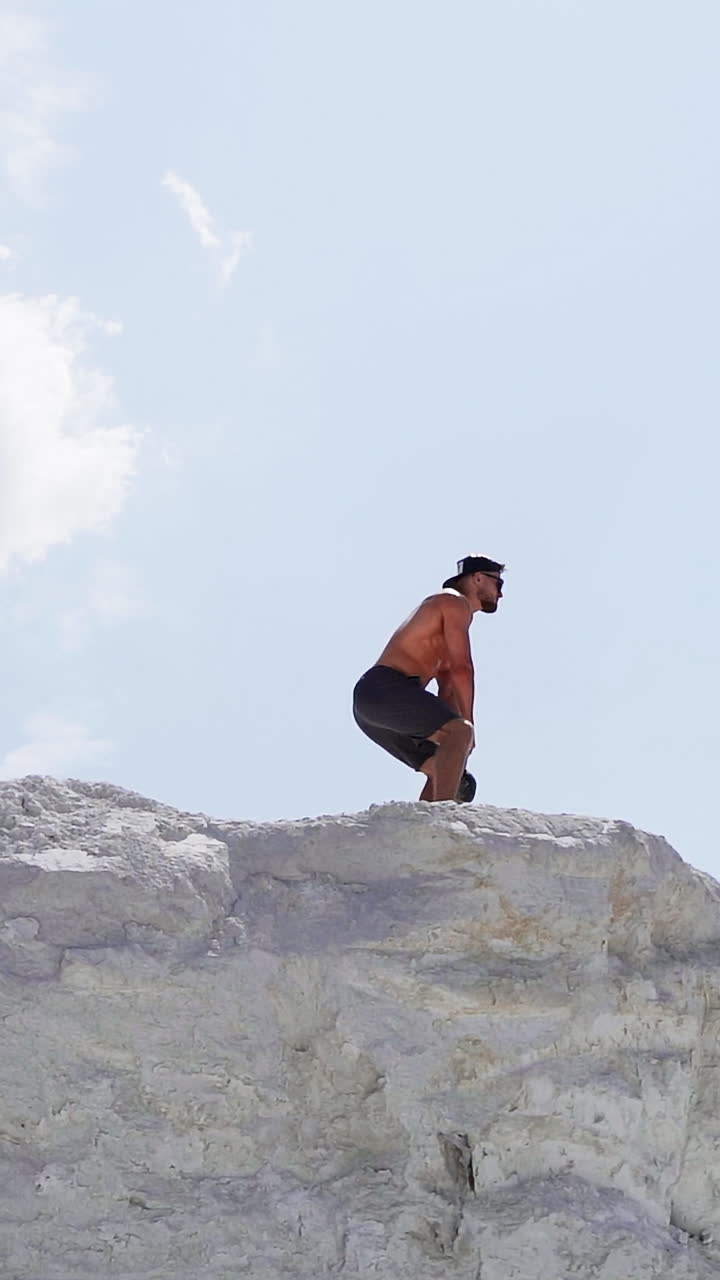 Side view of a strong man lifting kettlebell on white hill. Male athlete swinging kettlebell with two hands on the beautiful natural background outdoors. Vertical video
