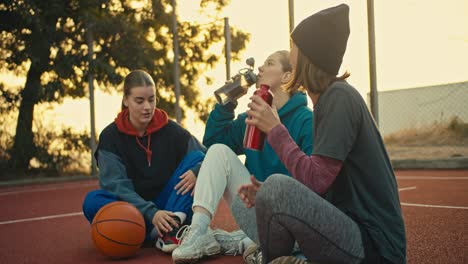 A-trio-of-blonde-girls-in-sportswear-drink-water-from-sports-bottles-and-chat-during-a-break-from-basketball-practice-near-an-orange-ball-on-a-red-street-court-at-sunrise