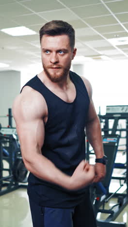 muscular bodybuilder showing strong biceps on arm. confident macho looks at his athletic body and trained muscles on the background of modern fitness club indoors. vertical video