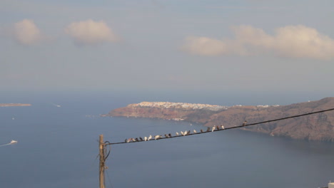 el pueblo de oia construido al borde de oscuros acantilados, visto desde lejos con palomas sentadas en un cable telefónico en primer plano