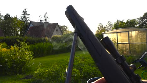 close up side view of a rifle after being shot, opened and it is smoking from the barrel where it needs to be reloaded. sunset evening at a sunny green grassy garden with a greenhouse in the back